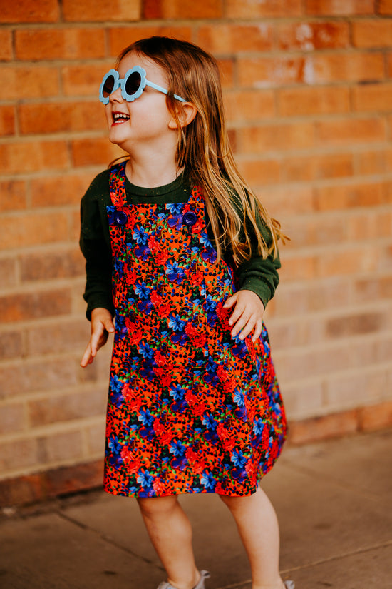 Girls handmade pinafore dress in autumn/winter floral orange and blue print. Pocket to front and button fastening straps. Available in ages 1-2 years up to 11-12 years